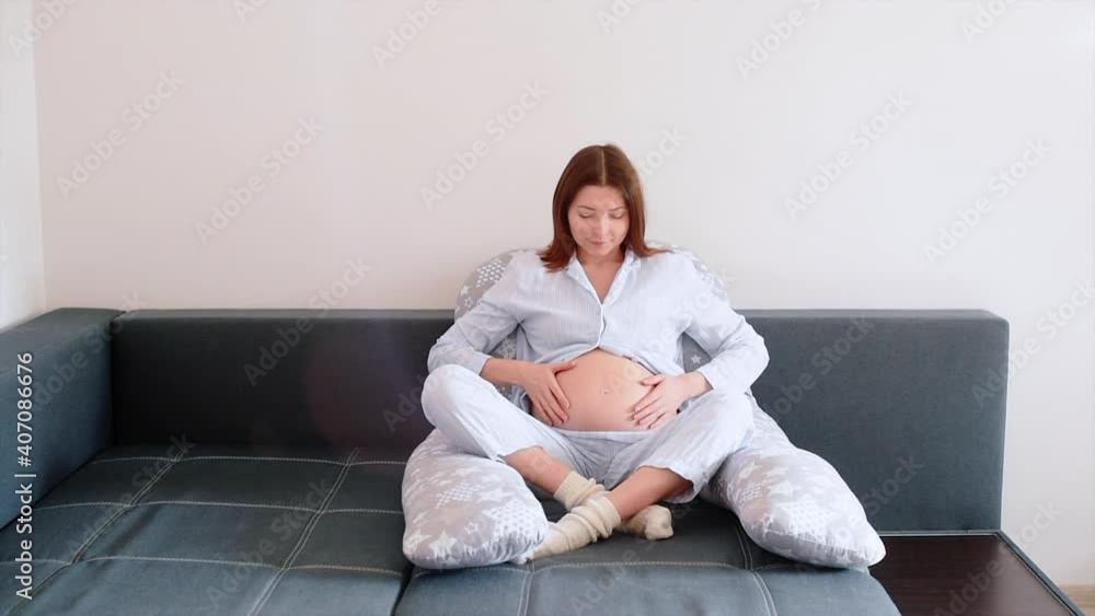 Happy pregnancy woman sitting on sofa at home in bedroom and holding hands on belly. Mom expecting baby. Concept of pregnancy, maternity, health care. Happy pregnancy woman sitting on sofa at home in bedroom and holding hands on belly. Mom expecting baby. Concept of pregnancy, maternity, health care.