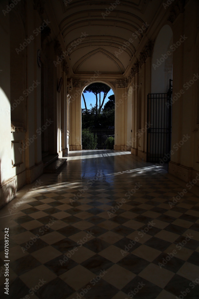 Roman church porch with a geometric patterned floor, partially illuminated by the sun and view of green pines and their reflection