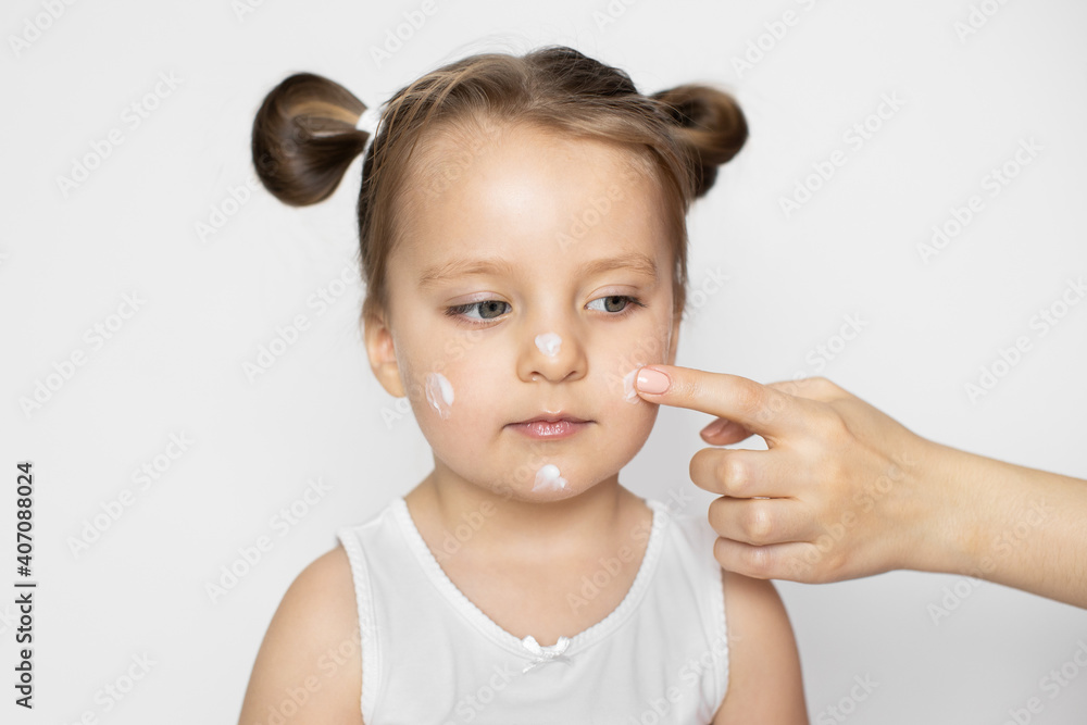 Skin care, hygiene, protection of baby's skin concept. Close up studio shot of mother's hand, applying lotion cream on face of the little beautiful baby girl after bathing to protect skin. Skin care, hygiene, protection of baby's skin concept. Close up studio shot of mother's hand, applying lotion cream on face of the little beautiful baby girl after bathing to protect skin.