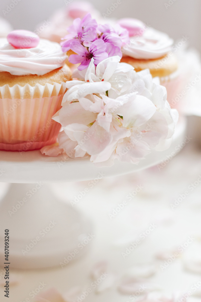 Vanilla cupcakes with white icing and candy topping served on cake stand with flowers and pink rose petals sprinkled for decoration