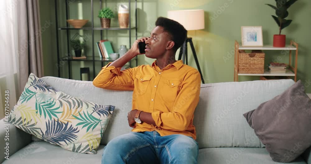 Camera approaching handsome young African american happy guy sitting on couch at home in cozy room alone talking on smartphone and smiling in positive mood. cellphone call, leisure concept Camera approaching handsome young African american happy guy sitting on couch at home in cozy room alone talking on smartphone and smiling in positive mood. cellphone call, leisure concept