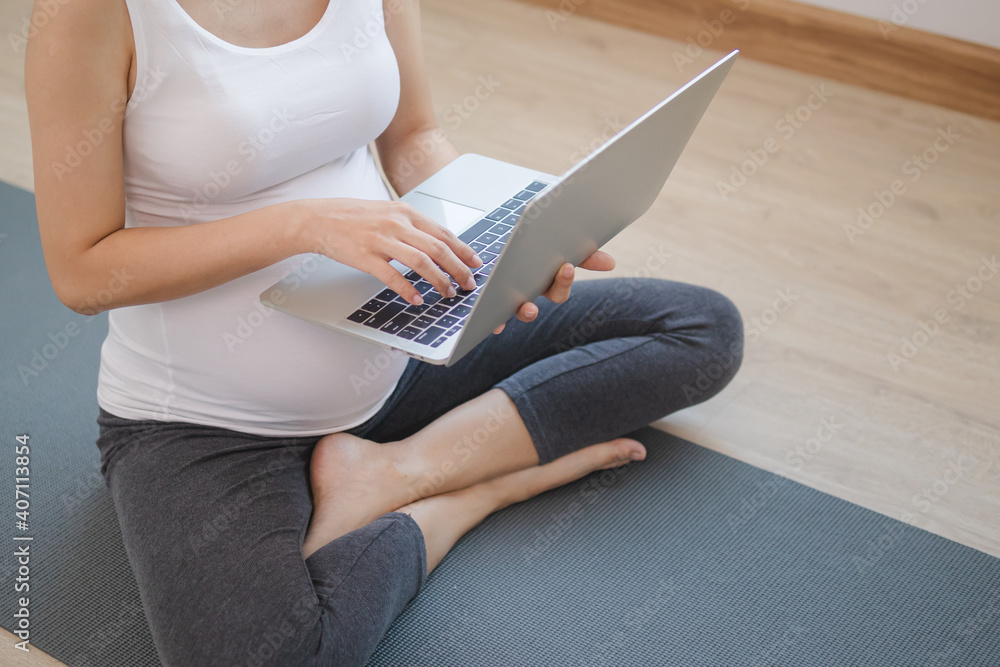 Fototapeta premium Calm pose young asian pregnant woman searching for yoga exercises for maternity while sitting on a mat via laptop at home.