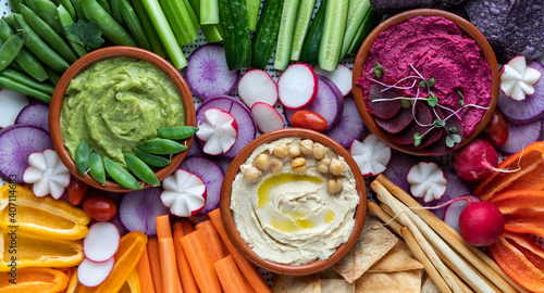 Top down close up view of three colourful hummus dips surrounded by various cut vegetables, pita bites and breadsticks.