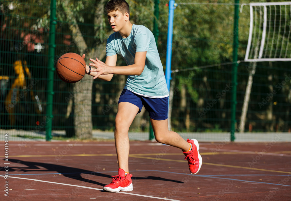 Naklejka premium Cute young boy plays basketball on street playground in summer. Teenager in green t shirt with orange basketball ball outside. Hobby, active lifestyle, sport activity for kids. 
