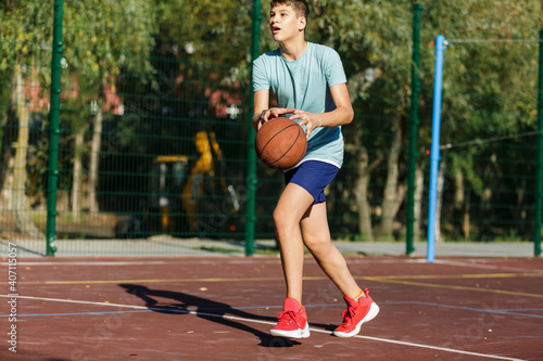 Cute young boy plays basketball on street playground in summer. Teenager in green t shirt with orange basketball ball outside. Hobby, active lifestyle, sport activity for kids. © Natali