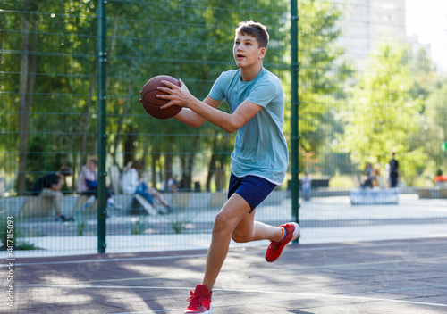 Cute young boy plays basketball on street playground in summer. Teenager in green t shirt with orange basketball ball outside. Hobby, active lifestyle, sport activity for kids.  © Natali