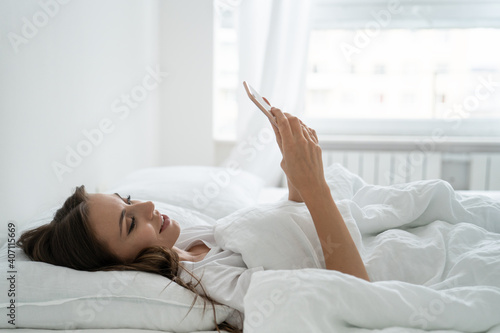 Girl enjoying lazy weekend morning. Smiling millennial young woman using mobile smart phone, lying and relaxing in white comfortable bed at home, chatting in social networks.