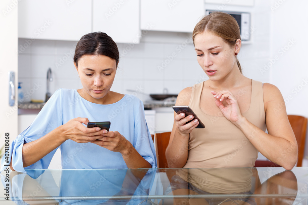 Obraz premium Two young girls having emotional conversation and looking at smartphone in kitchen