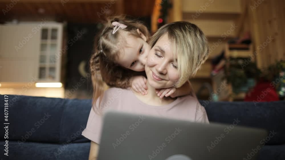 Distance job, family routine and parenthood concept. Young mom trying to work on laptop at home with her daughter cuddling kissing her in living room. Remote work during covid-19 pandemic, lockdown.