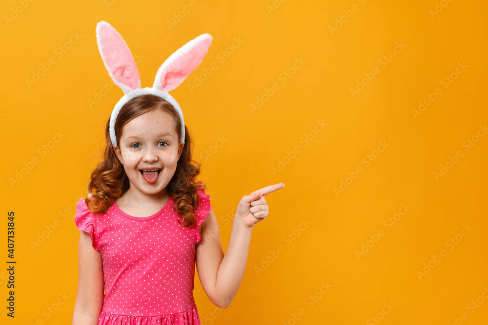 Cheerful happy cute little girl in Easter bunny ears on a yellow background. The child points with a finger to the copy space.