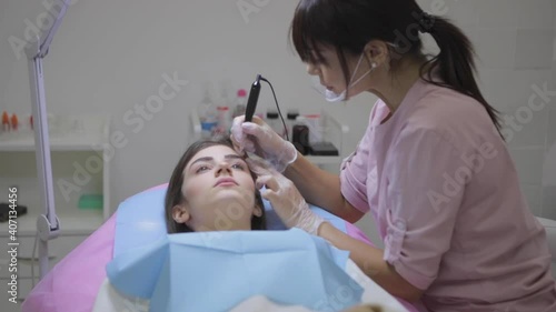Cosmetologist with plastic shied on face applies permanent makeup on young woman brows with tattoo machine in salon slow motion