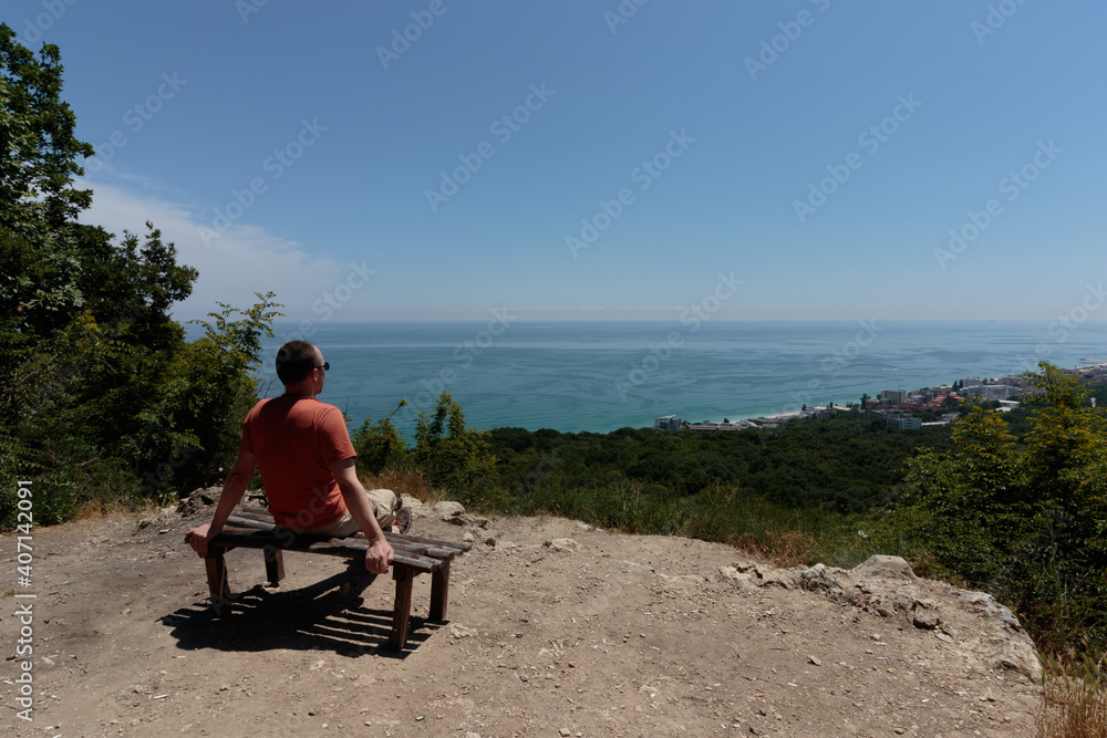 man sitting on a wooden bench and looking at the sea coast from the top of a hill