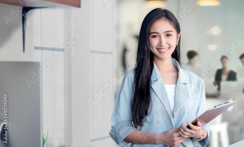 Obraz na plátně Portrait of young asian woman in casual wear holdng clipboard, smiling and looking at camera while standing in modern office room