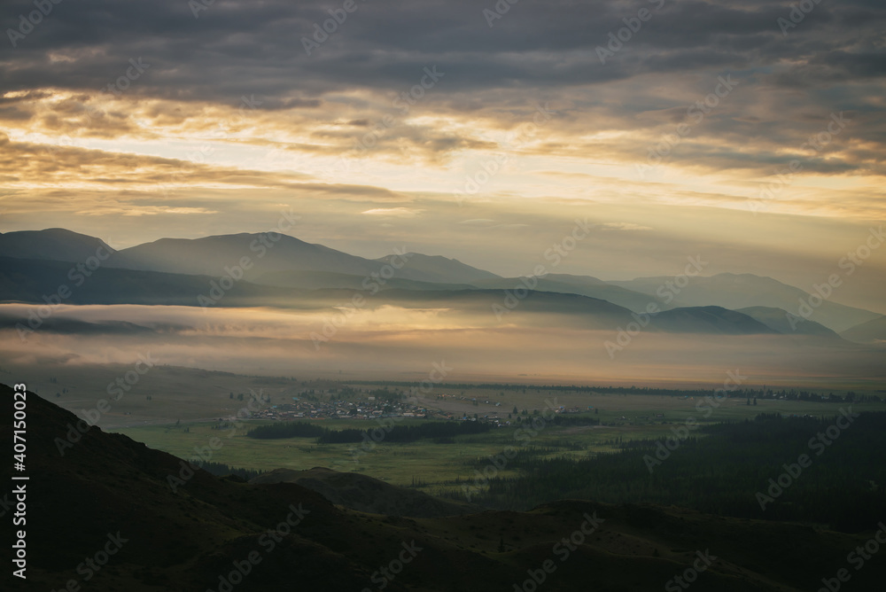 Scenic mountain landscape with golden low clouds above village in mountain valley among mountains silhouettes under dawn cloudy sky. Atmospheric alpine scenery of countryside in orange low clouds.