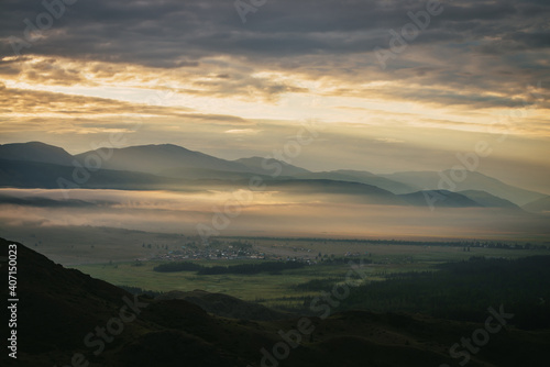 Scenic mountain landscape with golden low clouds above village in mountain valley among mountains silhouettes under dawn cloudy sky. Atmospheric alpine scenery of countryside in orange low clouds.