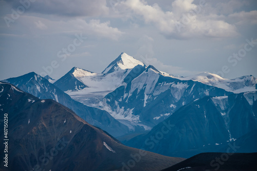 Awesome mountains landscape with big snowy mountain pinnacle in blue white colors under cloudy sky. Atmospheric highland scenery with high mountain wall with pointed top with snow under white clouds.