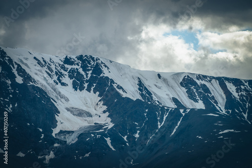 Dramatic mountains landscape with big snowy mountain ridge under cloudy sky. Dark atmospheric highland scenery with high mountain range in overcast weather. Awesome big mountain wall under gray clouds
