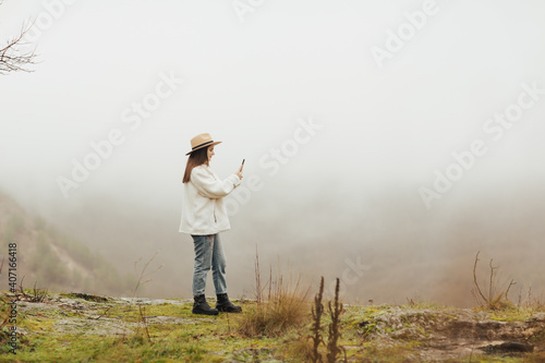Stylish blogger on a hill photographing a rocky destination, pine forest and foggy hills.