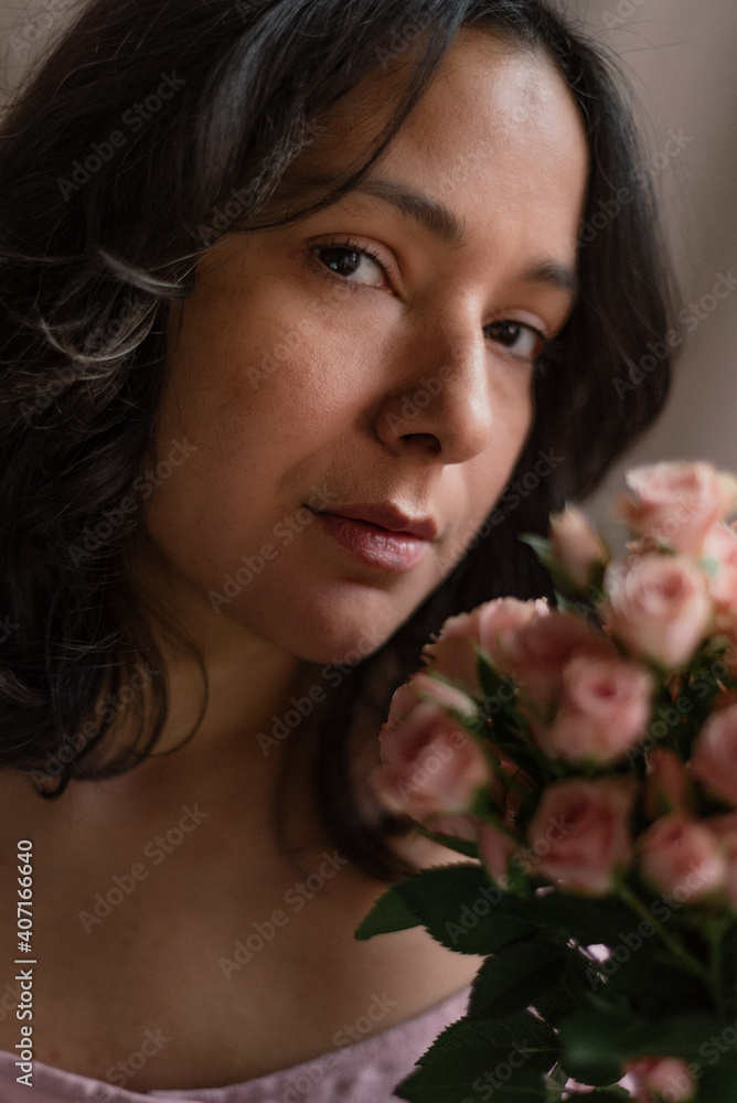 delicate young woman portrait with pink roses and pink dress and natural look vertical close up holding flowers bouquet. midlife fragile woman. confident femininity. dreamy expression and soft light.