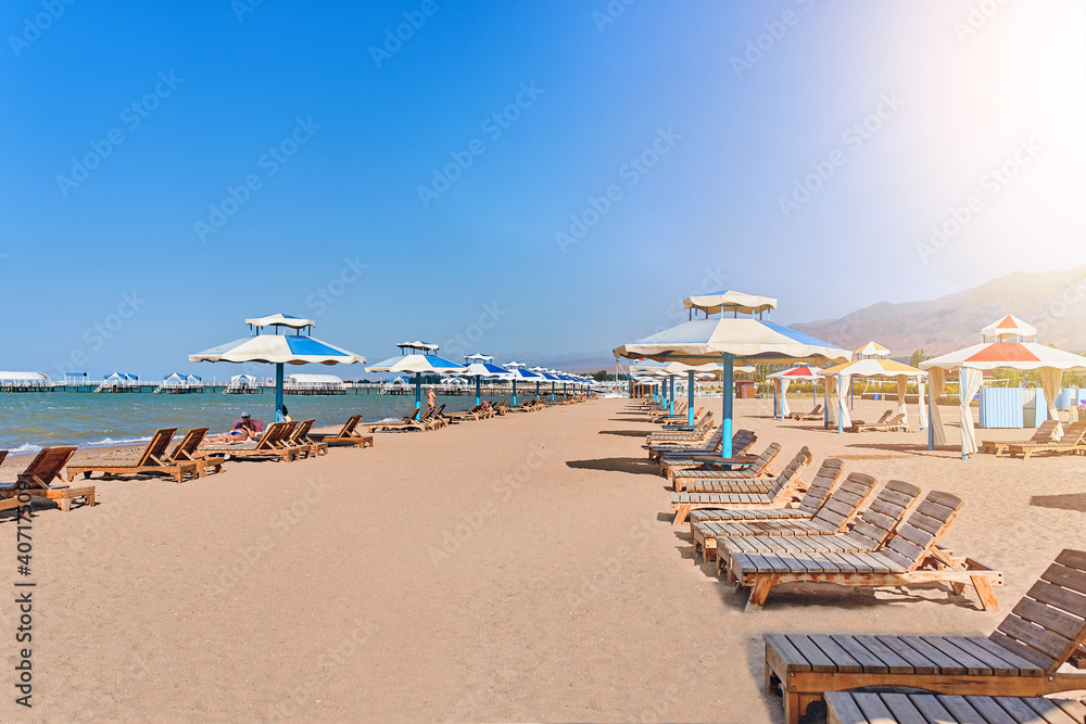 Obraz premium Sunbeds on beach. Umbrellas from the sun with seats on the lake. Wooden latticed sun beds. Beach at a boarding house or hotel. Vacation.