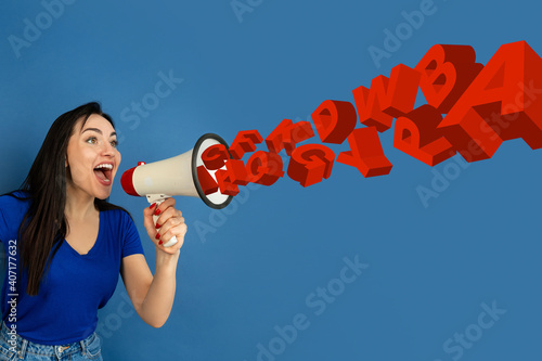 Young caucasian woman shouting with megaphone, loudspeaker on blue studio background. Sales, offer, business, cheering fun concept. Stream of letters flying from the inside of rupor. Copyspace for ad. photo