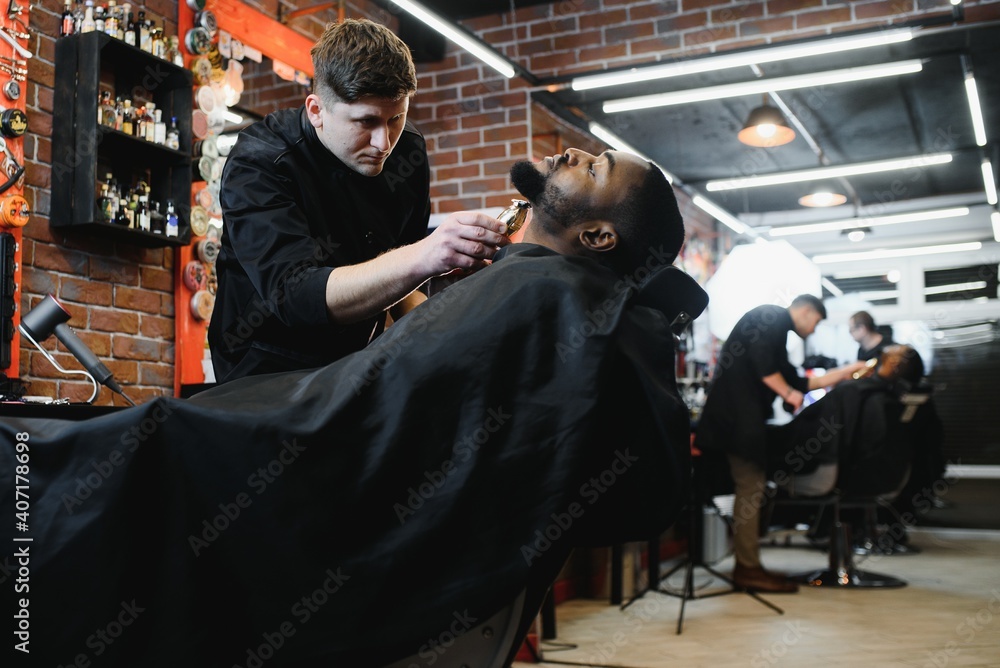 A barber is going through the electric cutting and shaving machine for the beard of an African-American Brazilian boy.