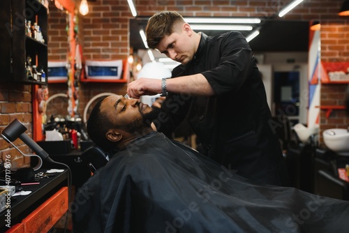 Side view of serious man with stylish modern haircut looking forward in barber shop. Hand of barber keeping straight razor and cutting trendy stripes on head of client.