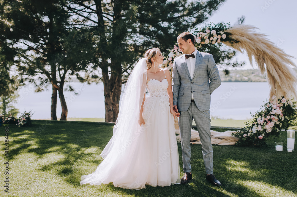Wedding portrait of smiling newlyweds. Stylish groom in a gray checkered suit and a cute blonde bride in a white lace dress stand holding hands in the forest against the background of the arch.