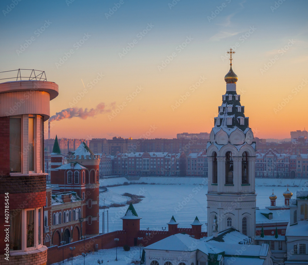 Fototapeta premium Winter frosty morning in Yoshkar-Ola city. Orange sunrise over the city. .Sunrise top view to the bell tower of the church and Kokshaga river embankment.