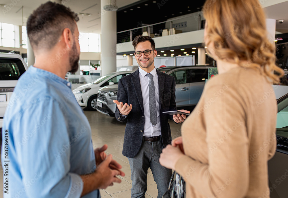 Fototapeta premium Young car salesman showing to mid-age couple new automobile at dealership salon.