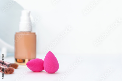Pink sponge and a bottle of beige cosmetic foundation  brushes for make-up and mirror on white table background  close-up. Makeup artist and woman accessories for perfect face skin  beauty  copyspace.