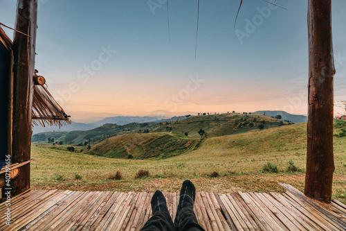 Tableau sur toile Legs of traveler relaxing in hut on green hill at sunrise
