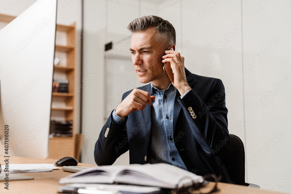 Displeased grey man talking on mobile phone while working with computer Displeased grey man talking on mobile phone while working with computer