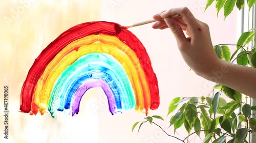 Little girl painting rainbow on window during Covid-19 quarantine at home. Stay at home social media campaign for coronavirus prevention, let's all be well, catch the rainbow photo