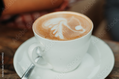Closeup shot of white cup filled with aromatic coffee with heart shape latte art foam on wood table near window with light shade on tabletop at cafe with spoon besides it 
