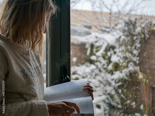 A shallow focus shot of a blonde woman reading a book and watching it snow in front of the window