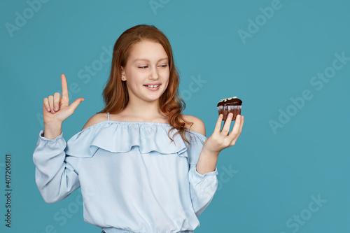 smiling redhead child girl is holding in hand cake and pointing up isolated over blue background