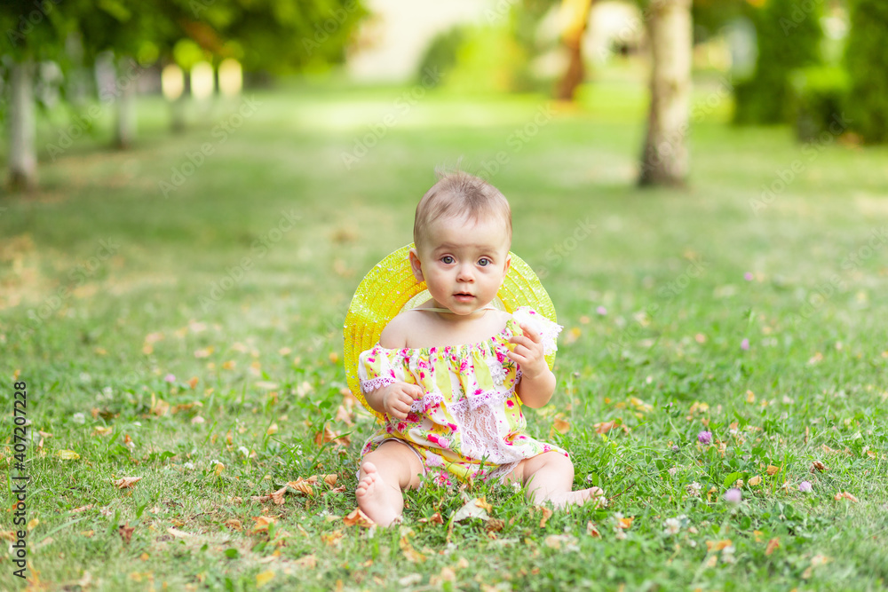 a small baby girl is sitting on the green grass in a yellow dress and hat and holding her hand to her ear, walking in the fresh air. Space for text