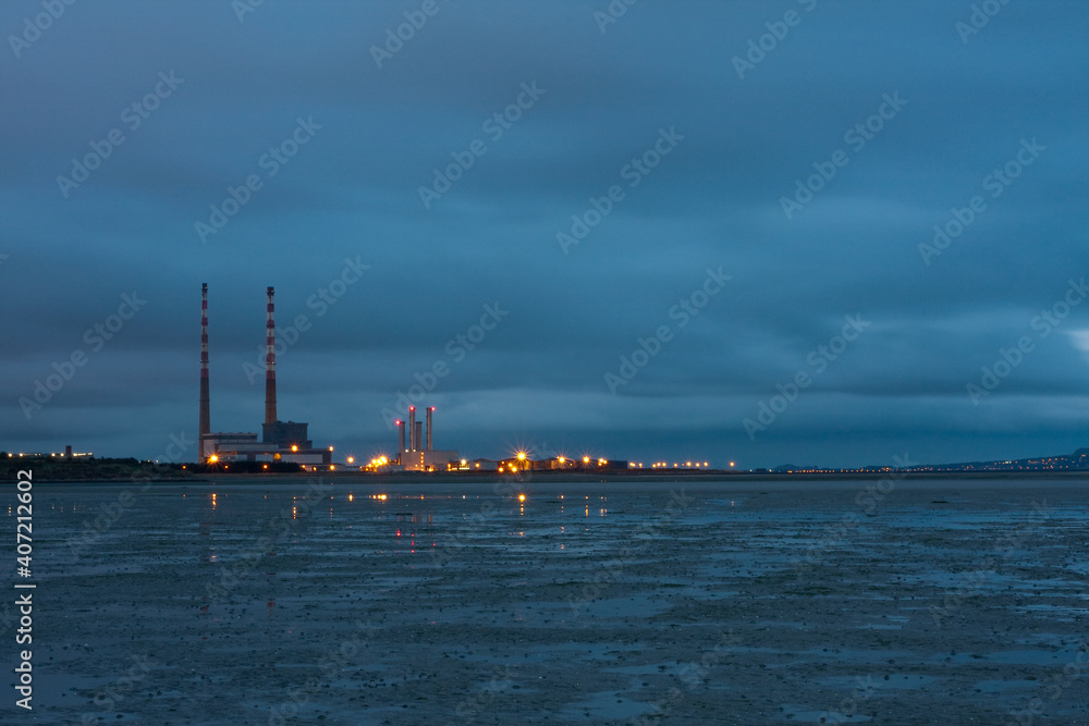 Landmark in Dublin Ireland. Poolbeg twin Chimneys at night. Dark blue cloudy sky, starburst lights and water reflected in pools with sea tide out. Decommissioned electricity generating station