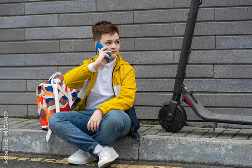 Modern teenager with backpack and electric scooter sitting on the sidewalk and talking on the phone
