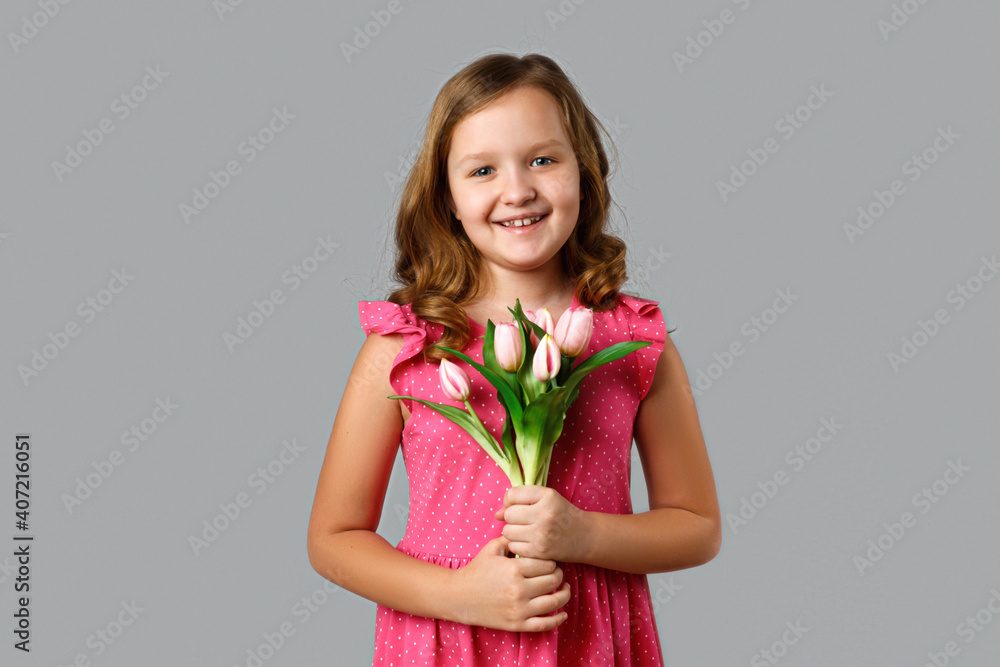 Cute happy little girl in a pink dress with polka dots on a gray background. The child is holding a bouquet of tulips. Cute happy little girl in a pink dress with polka dots on a gray background. The child is holding a bouquet of tulips.