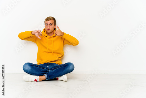 Young caucasian man sitting on the floor showing thumbs up and thumbs down, difficult choose concept © Asier