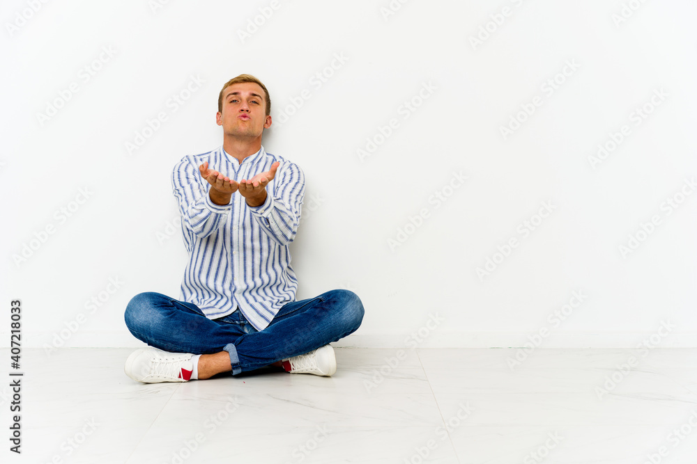 Young caucasian man sitting on the floor folding lips and holding palms to send air kiss. Young caucasian man sitting on the floor folding lips and holding palms to send air kiss.