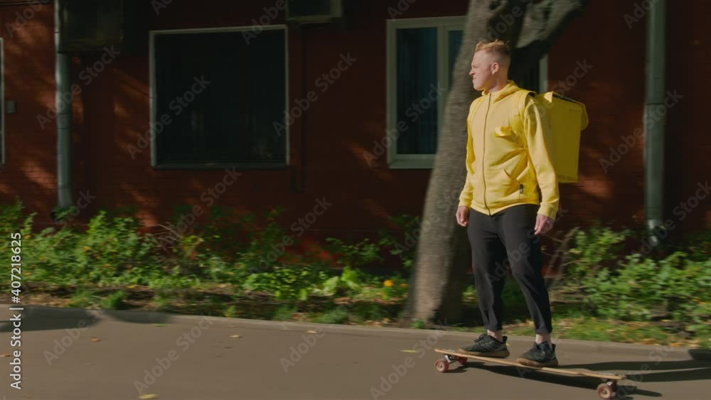Red residential building in background and parked cars in front of camera. Young courier guy, on longboard rides along shady street. Delivery person carrying online food order or takeout meals. Red residential building in background and parked cars in front of camera. Young courier guy, on longboard rides along shady street. Delivery person carrying online food order or takeout meals.