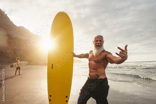 Happy fit senior having fun surfing at sunset time - Sporty bearded man training with surfboard on the beach - Elderly healthy people lifestyle and extreme sport concept photo