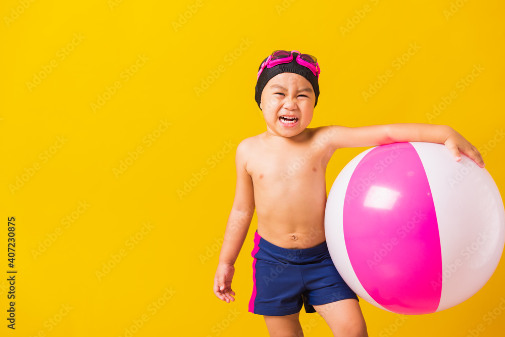 Summer vacation concept, Portrait Asian happy cute little child boy smiling in swimsuit hold beach ball, Kid having fun with inflatable ball in summer vacation, studio shot isolated yellow background