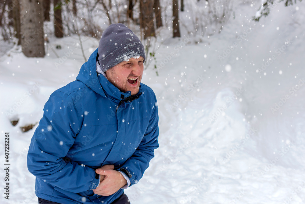 Young handsome caucasian man wearing cap and jacket on snowy weather with hand on stomach causing pain in stomach, feeling unwell. Photo of young man with stomach pain. Frustrated man with stomachache Young handsome caucasian man wearing cap and jacket on snowy weather with hand on stomach causing pain in stomach, feeling unwell. Photo of young man with stomach pain. Frustrated man with stomachache