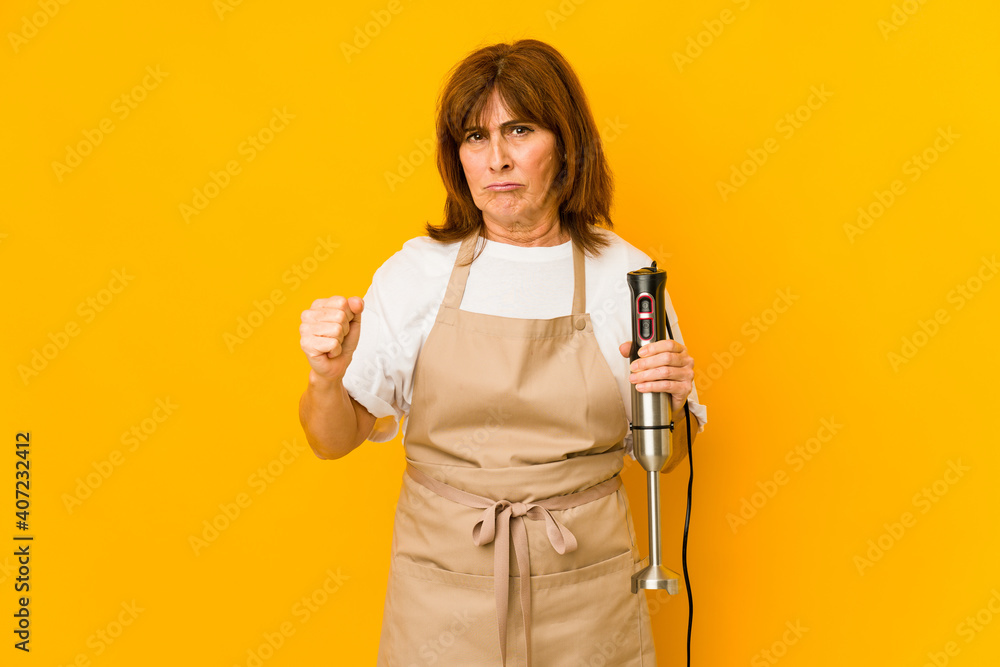 Middle age caucasian cook woman holding a mixer isolated showing fist to camera, aggressive facial expression. Middle age caucasian cook woman holding a mixer isolated showing fist to camera, aggressive facial expression.