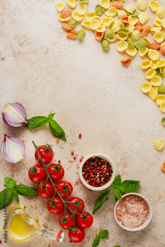 Raw orecchiette with parmesan cheese and tomatoes, basil, garlic and oil on light background with pepper on light brown background. Traditional ingredients for preparation of Italian pasta. Top view.