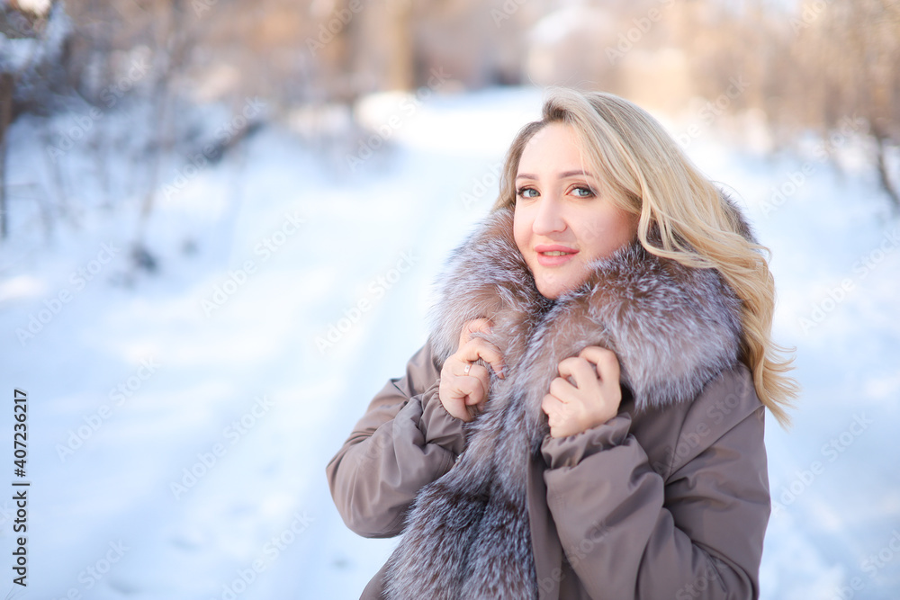 a blonde in a jacket with a fur collar stands sideways in a winter park